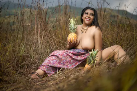 Smiling woman in a floral patterned skirt holding a pineapple in a grassy field during golden hour.