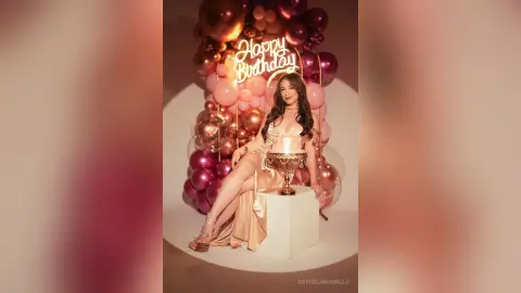 Woman in champagne satin dress celebrating birthday with rose gold balloons and neon sign in elegant setting.