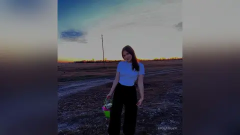 KailaDuch — Young woman in a white t-shirt and black trousers holding a flower basket in a rural field at sunset.