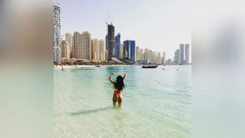 AnnaBanks stands in clear turquoise ocean water wearing a red bikini, facing away from the camera toward a distant city skyline.