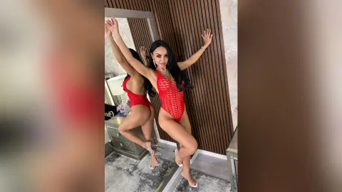 Glamorous woman in a red lace bodysuit posing in a modern luxury bedroom setting.