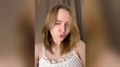 Blonde woman in a white lace camisole making a playful kissy face in a bright indoor setting.