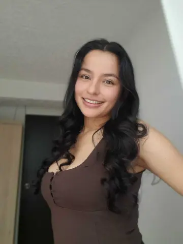 Smiling young woman with long wavy dark hair wearing a brown sleeveless top in a bright indoor setting.