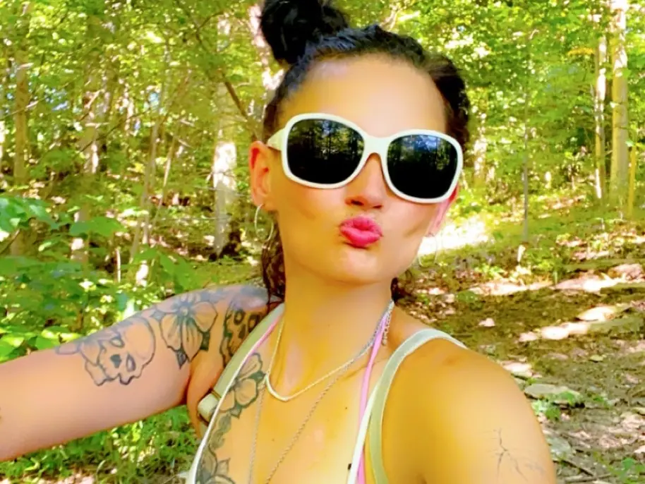 AnitaDickie — Woman with tattoos and white sunglasses making a kissy face in a sunny green forest setting.