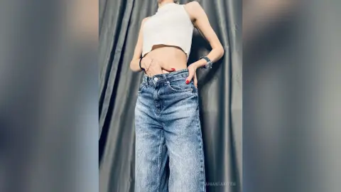 Young woman in a white crop top and high-waisted blue jeans posing against a grey curtain backdrop.