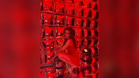 Glamorous woman in a red dress posing against a reflective silver textured wall under moody red lighting.