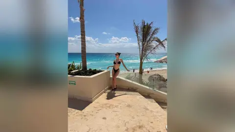Slim woman in a black bikini standing on a sunny tropical beach terrace near palm trees and turquoise ocean water.