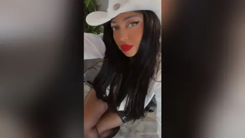 Glamorous woman with long dark hair and red lipstick wearing a white cowboy hat in a cozy indoor setting.