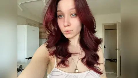 Young woman with wavy red hair wearing a white tank top in a bright indoor setting.