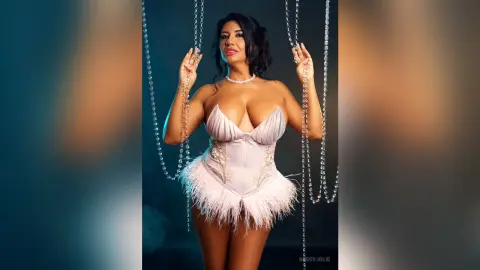 MissyJolie poses in a white feathered corset while holding long strands of silver beads against a dark background.