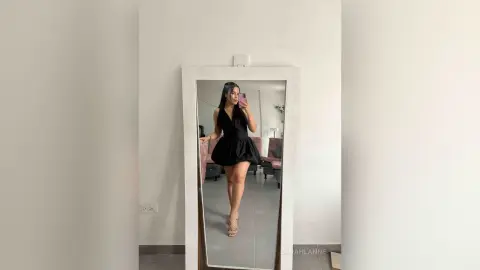 Young woman in a chic black mini dress taking a mirror selfie in a bright, minimalist room.