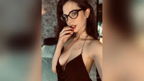Young woman with glasses in a black slip dress posing indoors with a confident and moody expression.