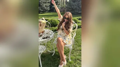 Cheerful woman in a floral summer dress sitting at a white garden table on a sunny lawn.