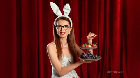 Smiling woman in white lace outfit and bunny ears holding a tray of grapes against red velvet curtains.