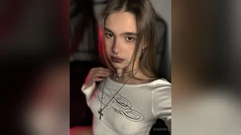 Young woman with long hair wearing a white graphic t-shirt and silver necklaces in a moody, dim indoor setting.