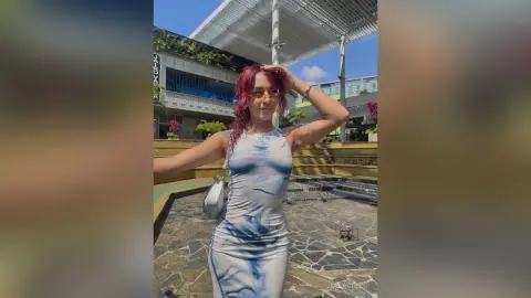 MilaGreco poses outdoors in a tight, white and blue tie-dye tank dress with her red hair styled down and sunglasses on.
