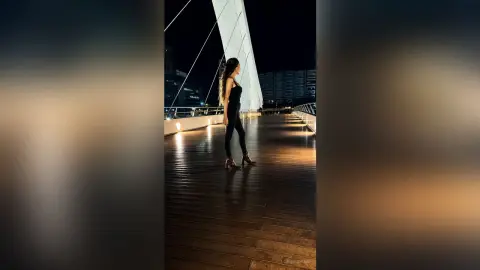 Stylish woman in a black jumpsuit standing on a wooden pier at night against a city skyline backdrop.