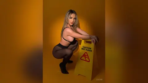 Lucine poses in a squatting position wearing a black bra and sheer black tights against a solid yellow background, leaning on a yellow caution sign.