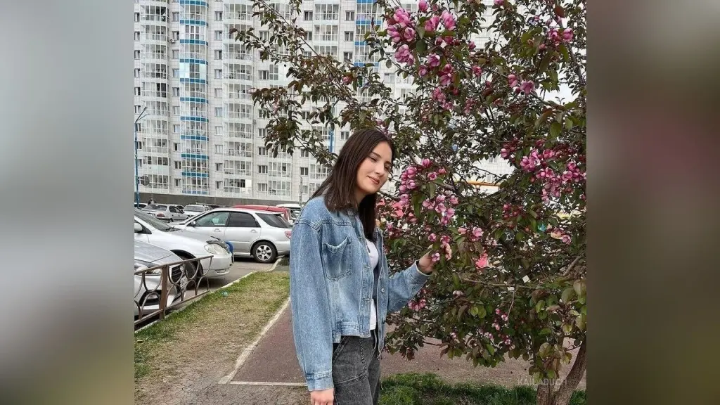 KailaDuch — Young woman in a denim jacket standing by a blooming pink flowering tree near city apartment buildings.
