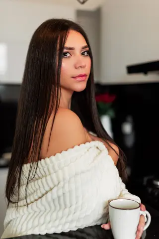 Young woman with long dark hair wearing a cozy white ribbed sweater holding a mug in a warm indoor setting.