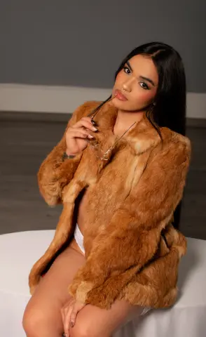 Glamorous woman in a brown faux fur coat sitting on a white stool against a grey studio background.