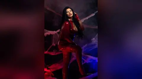 MissyJolie poses in a shiny red latex bodysuit and matching thigh-high boots against a dark, smoky background.
