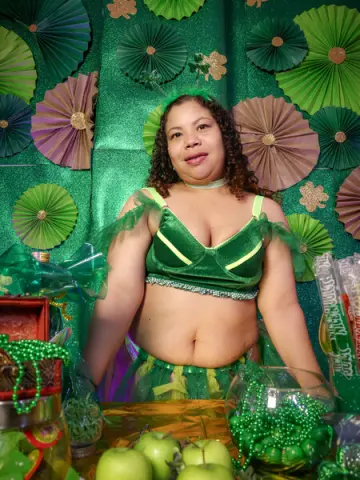 Smiling woman in a green velvet leotard posing before a festive green paper fan backdrop.