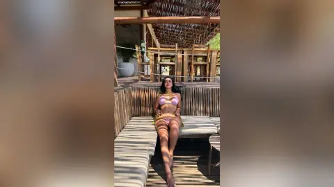 Young woman in a pink bikini relaxing on a wooden lounge chair at a sunny tropical beach resort.