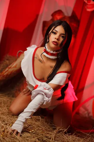 EmilyyJhonson wears a white and red maid outfit while posing on hay in front of red curtains.