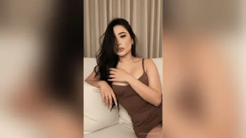 Elegant woman in a brown sleeveless dress posing confidently on a white sofa in a warm indoor setting.