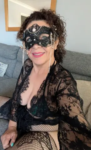 Smiling woman in a black lace robe and masquerade mask sitting on a grey sofa in a cozy indoor setting.