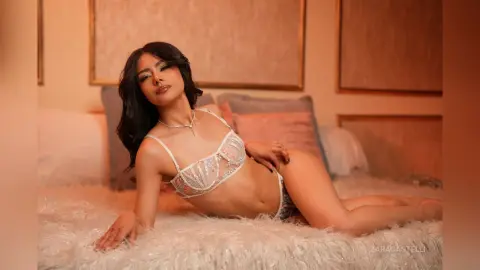 ZaraCastelli lies on a white furry bed wearing a white lace bra and matching panties, posing with her hand on her hip.