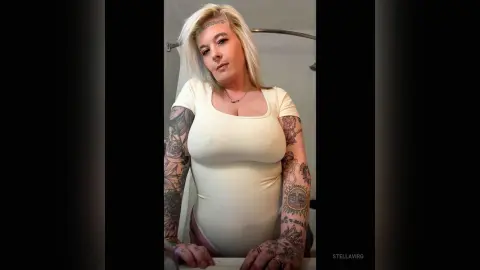 Tattooed woman in a white bodysuit posing in a bright bathroom setting.