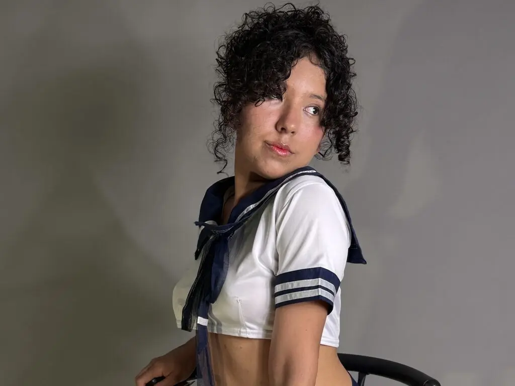 Young woman with curly hair wearing a white and navy sailor school uniform against a grey studio background.