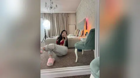 AriaVex sits on a carpeted floor in a bedroom, wearing a red bra and grey sweatpants while taking a mirror selfie.