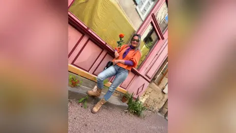 Stylish woman in orange top and blue jeans posing outside a pink building with a cheerful summer vibe.