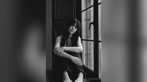 MintaTukes — MintaTukes sits by a window in a dark tank top and shorts, looking at the camera in a black and white photo.