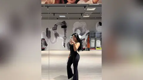 NicoleBella poses in a black sleeveless bodysuit while standing in a bright, modern gym setting.