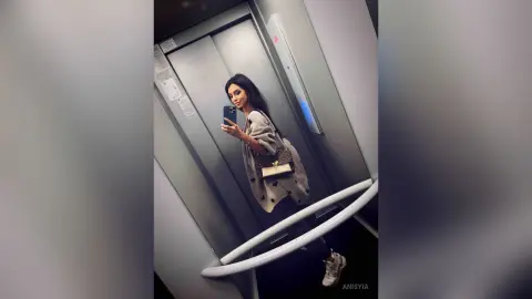 Stylish woman in a grey sweater taking a mirror selfie in a modern elevator.