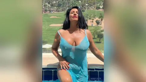 MissyJolie wears a sheer blue slip while posing outdoors against a green lawn and palm trees.