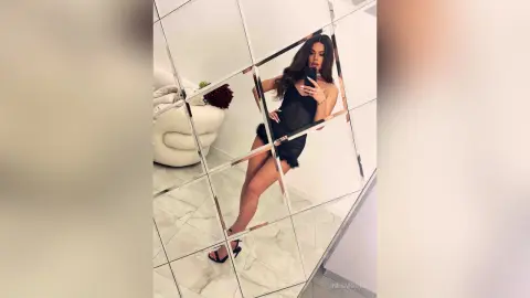 Glamorous woman in a chic black mini dress taking a mirror selfie in a bright, modern white room.
