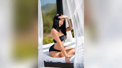 Beautiful brunette woman in a black bikini relaxing on a white canopy bed at a sunny outdoor resort.