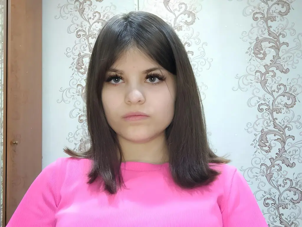 Young woman with long brown hair wearing a bright pink ribbed shirt against a floral patterned wallpaper background.