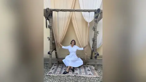 Woman in a white summer dress sitting on a wooden swing decorated with cream drapes in a bohemian setting.