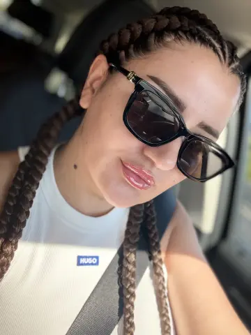 Smiling woman with long brown braids and black sunglasses wearing a white Hugo tank top in a car.