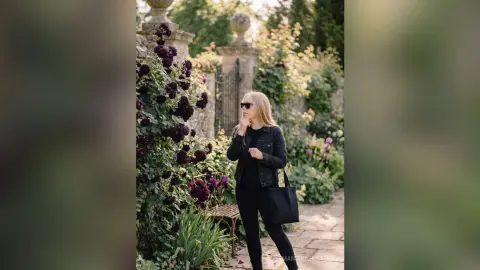 Blonde woman in a black denim jacket and sunglasses standing in a lush garden with purple flowers.