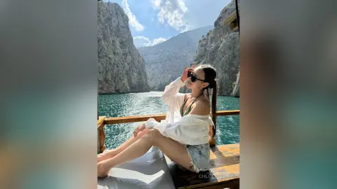 ChloeTeles sits on a wooden boat deck in a white off-the-shoulder top and denim shorts, posing against a backdrop of steep mountains and blue water.