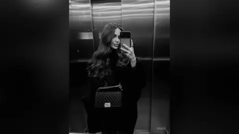 Stylish woman in a black outfit holding a designer quilted handbag in a modern elevator mirror selfie.