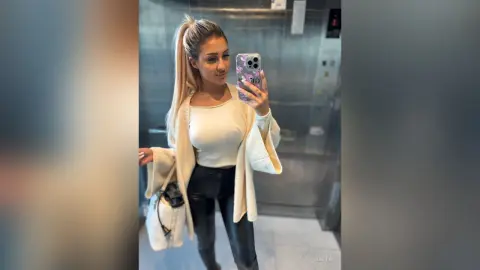Anita poses for a mirror selfie in an elevator wearing a white crop top and black leather leggings.