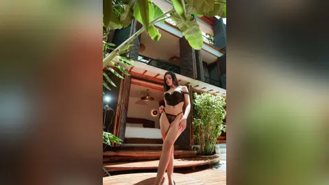 Slim woman in black lingerie posing confidently on a tropical wooden balcony near lush green plants.